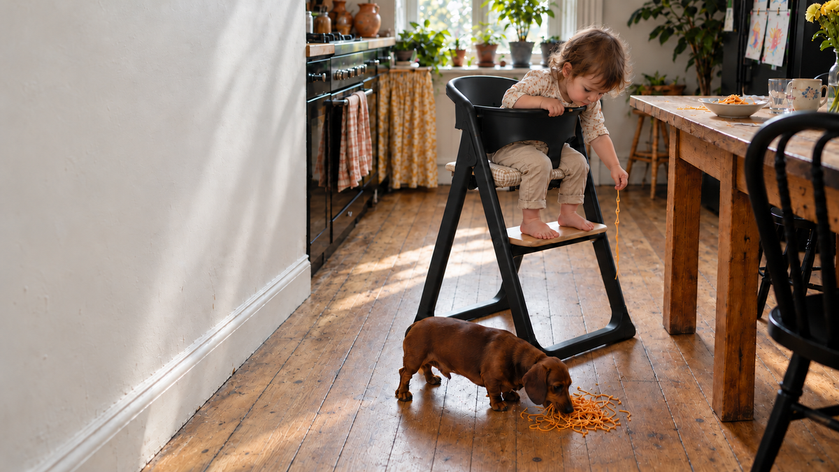 Child seated beside a kitchen table in the black Alto highchair with a houndstooth seat cushion and feet supported on the wooden footplate, dropping pasta to a dog below.