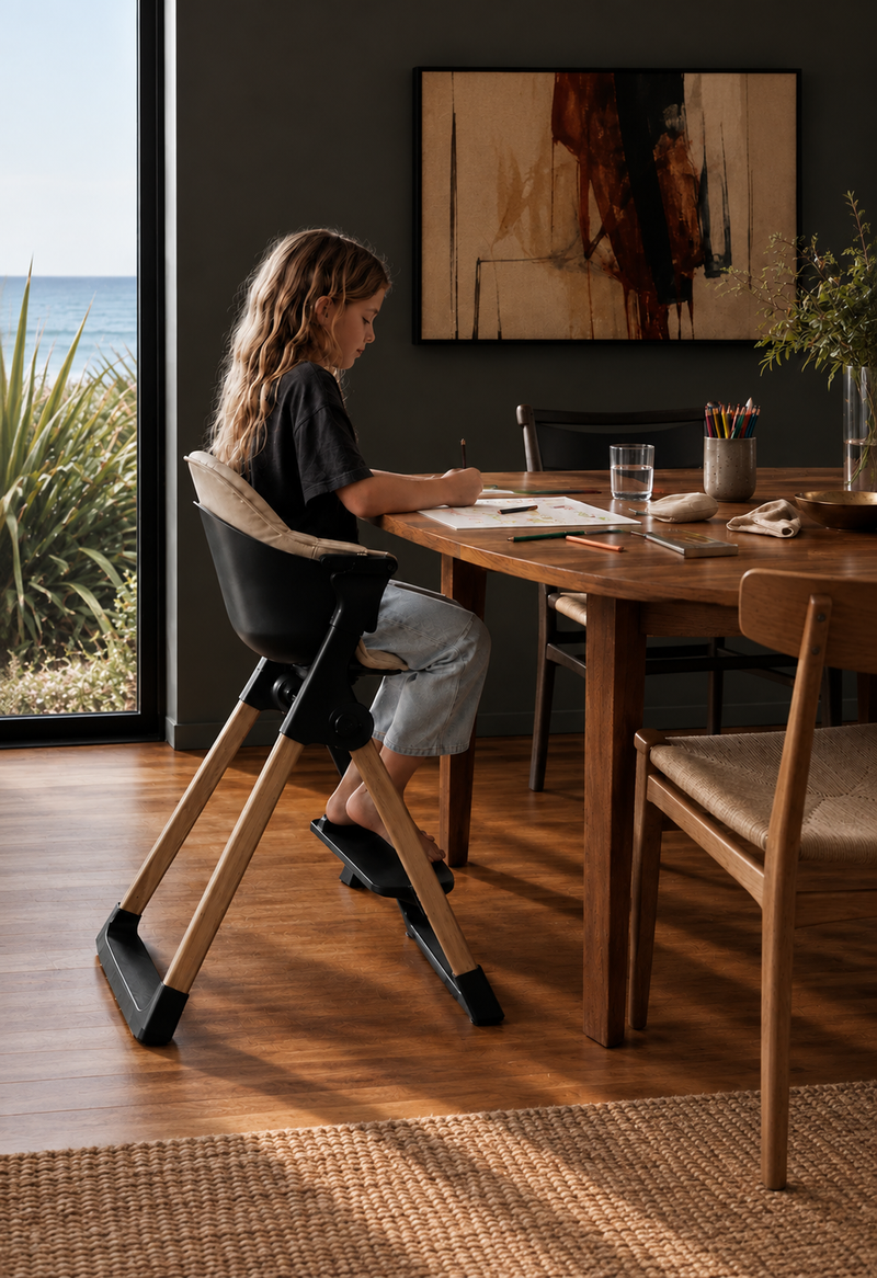 Older child writing at a table in the black Luna chair with natural wooden legs and the tray and safety rail removed.